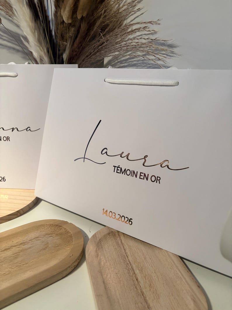 May include: White gift bag with a gold-colored script that reads "Laura" and "T&eacute;moin en Or". The date "14.03.2026" is printed in gold. The bag has a white cord handle.