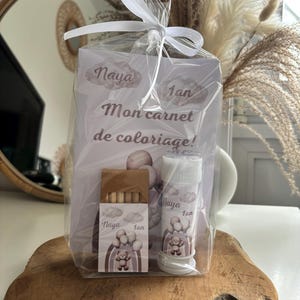 Peut inclure: Un coffret cadeau pour le premier anniversaire d'un enfant, enveloppé dans du cellophane transparent et noué avec un ruban blanc. L'ensemble comprend un cahier de coloriage, une petite boîte de crayons et un flacon de bulles. L'emballage présente un ours en peluche et un arc-en-ciel.