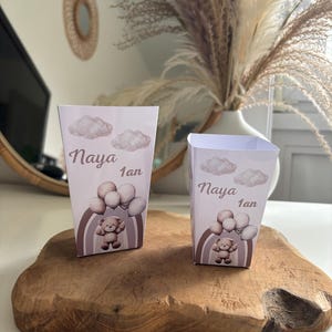 Puede incluir: Dos cajas de regalo de fiesta rosa y blancas con un diseño de osito de peluche, arcoíris y nubes. Las cajas dicen "Naya 1an".