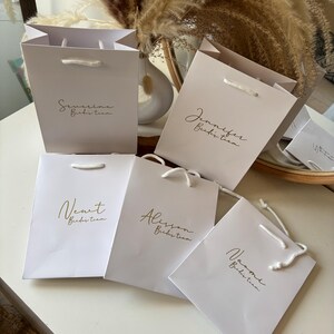 May include: A collection of white and light pink gift bags with gold lettering. Each bag has a name and the words "Bride Team" printed on it. The bags have white rope handles and are arranged on a white surface.