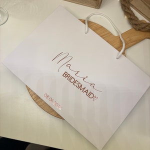 May include: A white gift bag with the words "Maria Bridesmaid" in a copper-colored script. The date "08.06.2024" is printed below. The bag has a white cord handle.