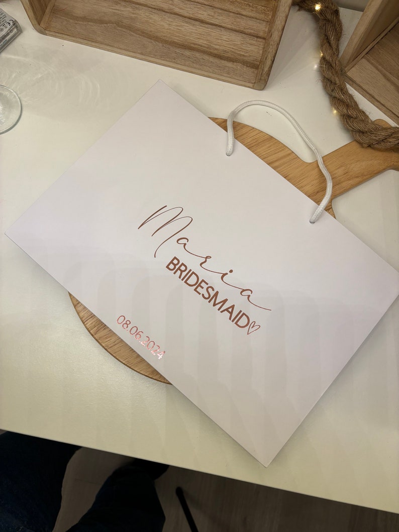 May include: A white gift bag with the words "Maria Bridesmaid" in a copper-colored script. The date "08.06.2024" is printed below. The bag has a white cord handle.