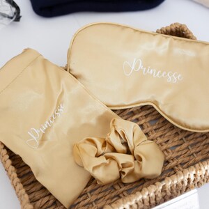 Könnte beinhalten: Ein goldfarbenes Schlafset mit Augenmaske, Scrunchie und Beutel, alle mit dem Wort "Princesse" in weißer Schrift bestickt. Die Artikel sind in einem Weidenkorb angeordnet.