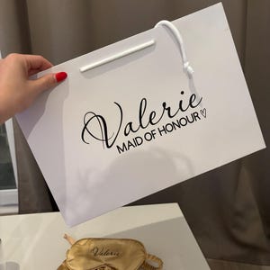 May include: A white gift bag with the name "Valerie" and "MAID OF HONOUR" printed in black script. A gold sleep mask, scrunchie, and pouch are also visible. The items are likely part of a bridal party gift set.