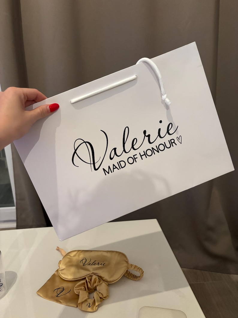 May include: A white gift bag with the name "Valerie" and "MAID OF HONOUR" printed in black script. A gold sleep mask, scrunchie, and pouch are also visible. The items are likely part of a bridal party gift set.