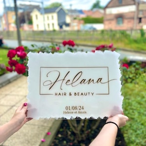 Może przedstawiać: Prostokątny szyld z matowego akrylu o szorstkich, teksturowanych krawędziach. Szyld zawiera napis "Helana HAIR & BEAUTY" brązową czcionką w prostokątnej ramce, a poniżej datę "01/08/24" oraz tekst "Helene et Anais".