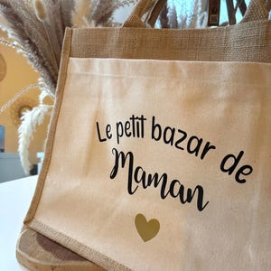 Peut inclure: Un sac fourre-tout beige avec une bordure et des poignées en toile de jute naturelle. Le sac porte l'inscription "Le petit bazar de Maman" en écriture noire, ainsi qu'un cœur doré. Le sac est conçu pour transporter des articles.