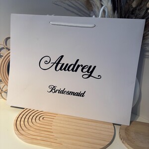May include: A white gift bag with the name "Audrey" in elegant script and the word "Bridesmaid" printed below. The bag has a white cord handle and sits on a light-colored wooden tray with a curved design.