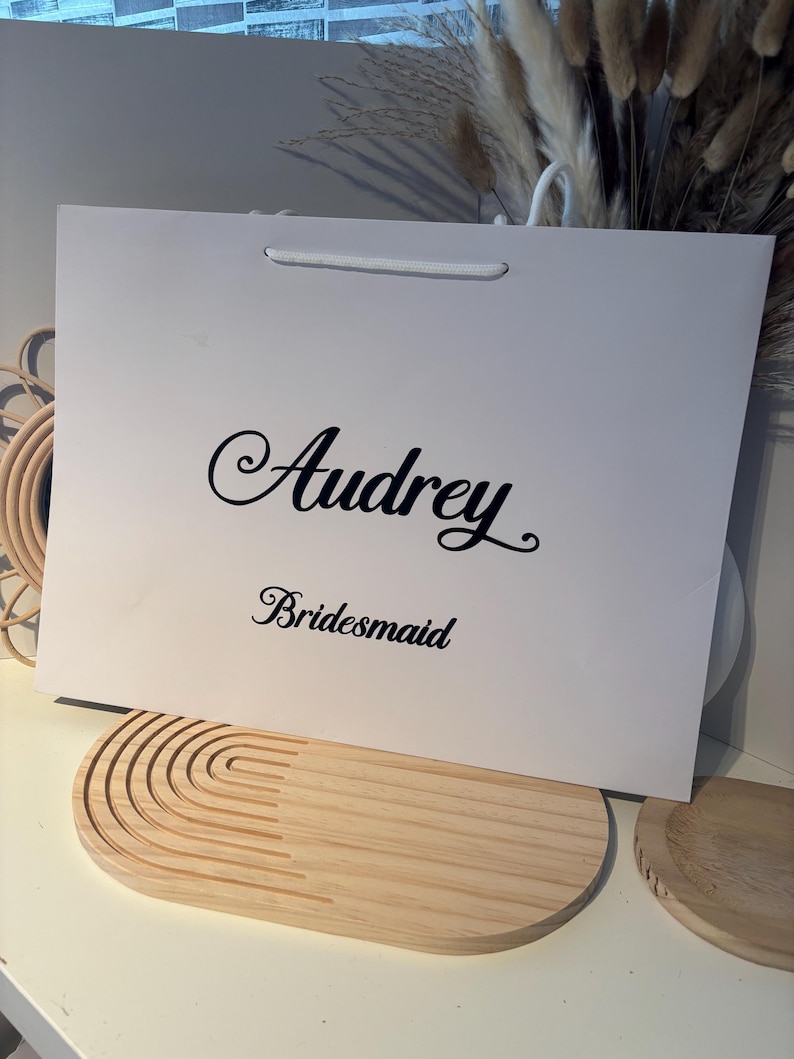 May include: A white gift bag with the name "Audrey" in elegant script and the word "Bridesmaid" printed below. The bag has a white cord handle and sits on a light-colored wooden tray with a curved design.