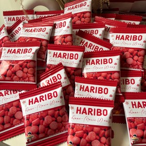 Könnte beinhalten: Eine Ansammlung von roten und weißen HARIBO-Bonbontüten. Jede Tüte trägt das HARIBO-Logo und den Text "Anne-Marie 60 ans". Die Tüten sind mit roten Süßigkeiten gefüllt und gestapelt.