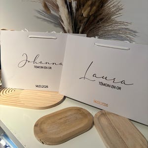 May include: Two white gift bags with the names "Johanna" and "Laura" in cursive script. The text "T&eacute;moin en Or" and the date "14.03.2026" are printed below the names. Two wooden oval trays are in the foreground.