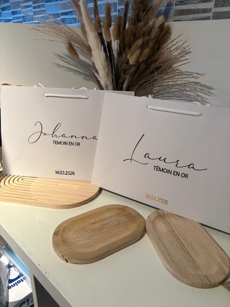 May include: Two white gift bags with the names "Johanna" and "Laura" in cursive script. The text "T&eacute;moin en Or" and the date "14.03.2026" are printed below the names. Two wooden oval trays are in the foreground.