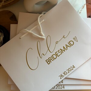 May include: White gift bag with gold lettering that reads "Chloe BRIDESMAID" and the date "26.10.2024". The bag has white rope handles and is stacked with other similar bags. The background includes a white ceramic sculpture and dried plants.