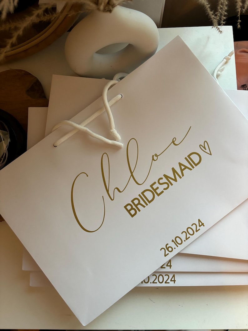 May include: White gift bag with gold lettering that reads "Chloe BRIDESMAID" and the date "26.10.2024". The bag has white rope handles and is stacked with other similar bags. The background includes a white ceramic sculpture and dried plants.