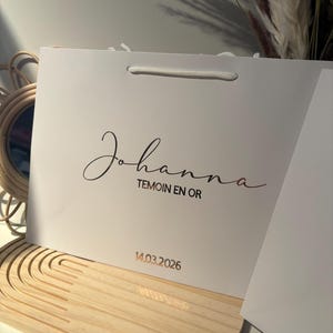 May include: A white gift bag with the name "Johanna" in elegant script, along with the text "TEMOIN EN OR" and the date "14.03.2026" in a gold color. The bag has a white rope handle.