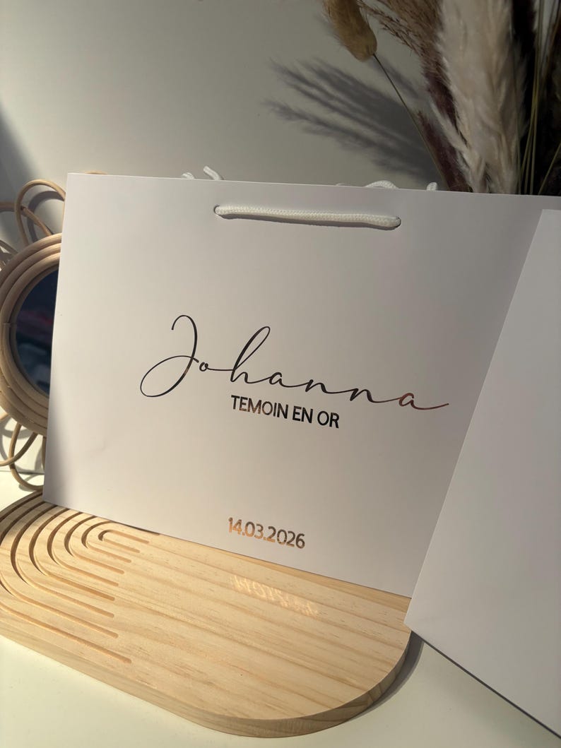 May include: A white gift bag with the name "Johanna" in elegant script, along with the text "TEMOIN EN OR" and the date "14.03.2026" in a gold color. The bag has a white rope handle.