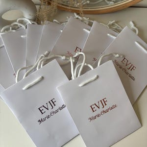 May include: A collection of white paper gift bags with white rope handles. Each bag has the text "EVJF Marie-Charlotte" printed in a burgundy color. The bags are arranged on a white surface.