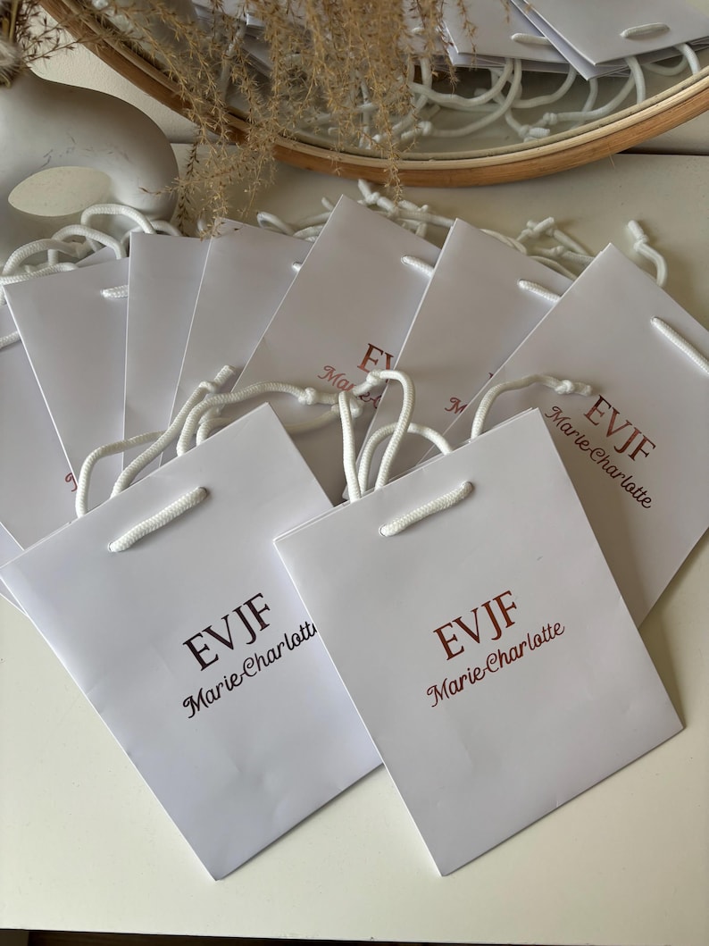May include: A collection of white paper gift bags with white rope handles. Each bag has the text "EVJF Marie-Charlotte" printed in a burgundy color. The bags are arranged on a white surface.