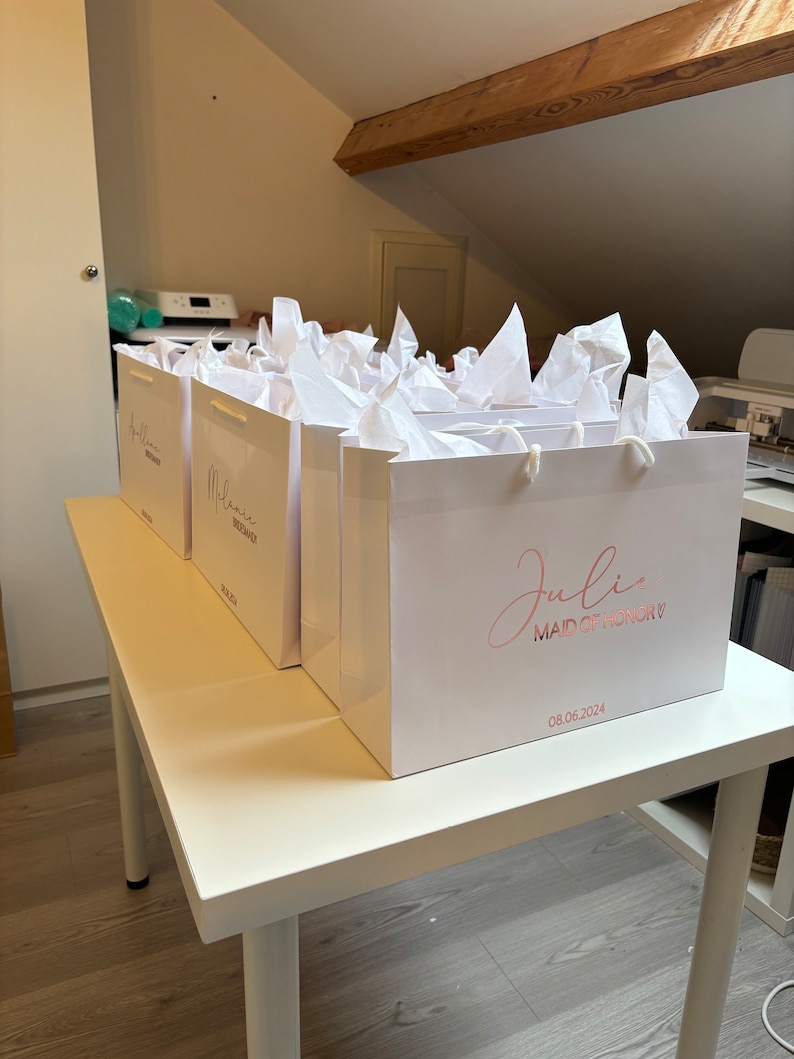 May include: White gift bags with tissue paper, each personalized with a name and date in rose gold lettering. One bag reads "Julie, Maid of Honor" with a date. The bags are on a white table.