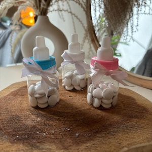 Puede incluir: Tres recipientes de plástico transparente con forma de biberón llenos de caramelos blancos. Las botellas tienen lazos de cinta azul, rosa y blanco.