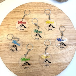 Op de afbeelding: Zes transparante acryl sleutelhangers met een bergontwerp en het woord "Merci" in verschillende kleuren. Elke sleutelhanger heeft ook het jaar "2024" erop gedrukt.