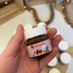 Peut inclure: Un petit pot en verre de pâte à tartiner au chocolat avec un couvercle blanc et une étiquette qui dit "Romain Candilier". L'étiquette présente également une illustration de dessin animé d'un couteau étalant la pâte à tartiner au chocolat sur du pain, un verre de lait et une noisette.