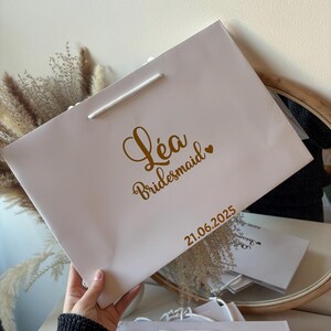 May include: A light pink gift bag with a white rope handle. The bag has gold text that reads "L&eacute;a Bridesmaid" with a heart, and the date "21.06.2025".