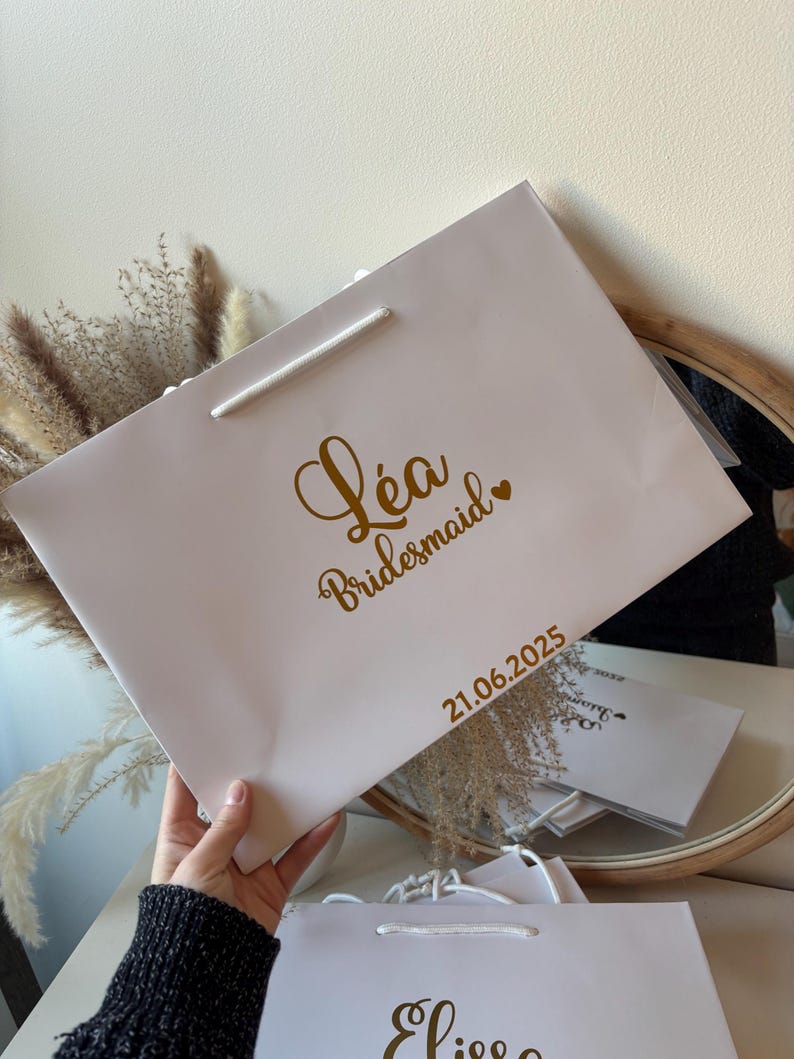 May include: A light pink gift bag with a white rope handle. The bag has gold text that reads "L&eacute;a Bridesmaid" with a heart, and the date "21.06.2025".