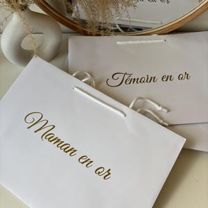 May include: White gift bags with gold lettering. One bag reads "Maman en or" and another reads "T&eacute;moin en or". The bags have white rope handles. A white vase and dried flowers are in the background.