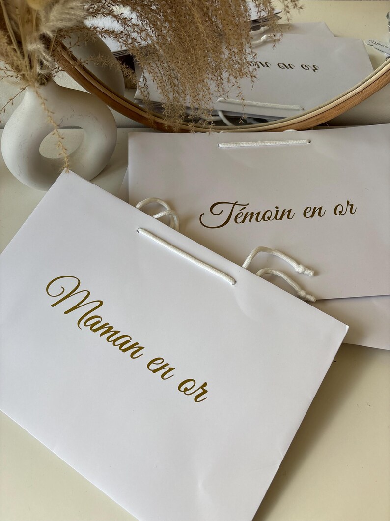 May include: White gift bags with gold lettering. One bag reads "Maman en or" and another reads "T&eacute;moin en or". The bags have white rope handles. A white vase and dried flowers are in the background.