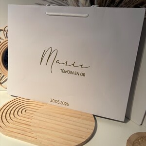 May include: A white gift bag with a gold script font that reads "Marie" and "T&eacute;moin en Or". The date "30.05.2026" is printed below. The bag has a white cord handle.
