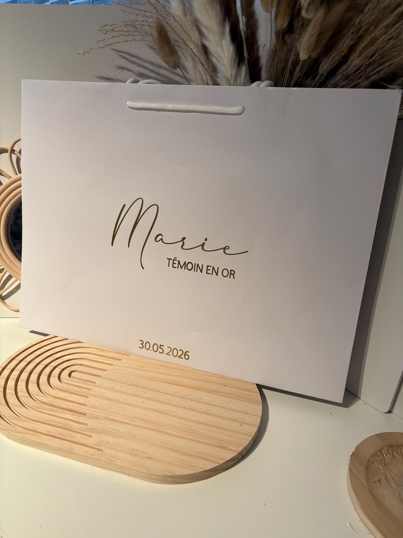 May include: A white gift bag with a gold script font that reads "Marie" and "T&eacute;moin en Or". The date "30.05.2026" is printed below. The bag has a white cord handle.