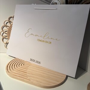 May include: A white gift bag with gold script text that reads "Emmeline T&Eacute;MOIN EN OR" and the date "30.05.2026". The bag has a white handle and is resting on a light wood decorative tray.