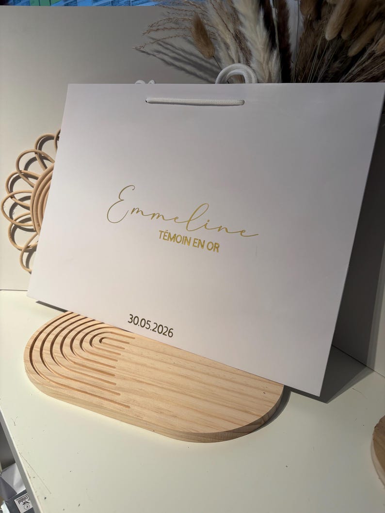 May include: A white gift bag with gold script text that reads "Emmeline T&Eacute;MOIN EN OR" and the date "30.05.2026". The bag has a white handle and is resting on a light wood decorative tray.