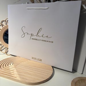 May include: A white gift bag with a gold script name "Sophie" and the text "DEMOISELLE D'HONNEUR EN OR". The date "30.05.2026" is printed below. The bag has a white cord handle.