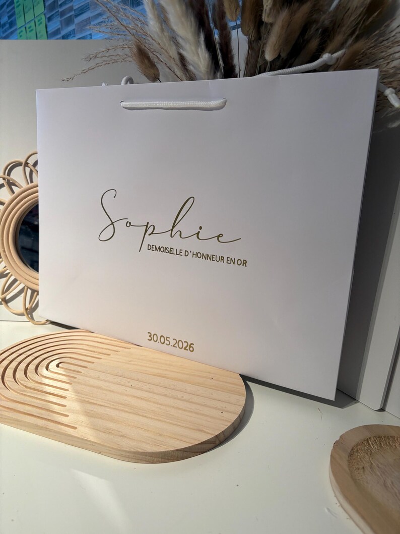 May include: A white gift bag with a gold script name "Sophie" and the text "DEMOISELLE D'HONNEUR EN OR". The date "30.05.2026" is printed below. The bag has a white cord handle.