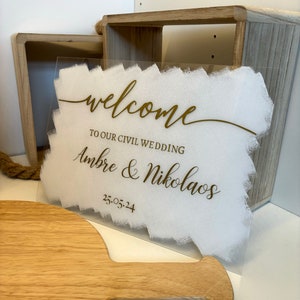 Könnte beinhalten: Ein klares Acrylschild mit weißem Hintergrund und goldenen Buchstaben. Das Schild lautet "Willkommen zu unserer standesamtlichen Hochzeit Ambre & Nikolaos 25.05.24".