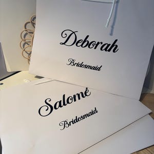 May include: Two white gift bags with black text. The top bag reads "Deborah" above "Bridesmaid" in a script font. The bottom bag reads "Salom&eacute;" above "Bridesmaid" in a script font. White string handles are visible.