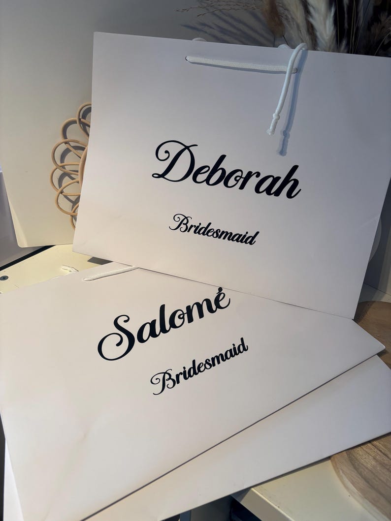 May include: Two white gift bags with black text. The top bag reads "Deborah" above "Bridesmaid" in a script font. The bottom bag reads "Salom&eacute;" above "Bridesmaid" in a script font. White string handles are visible.