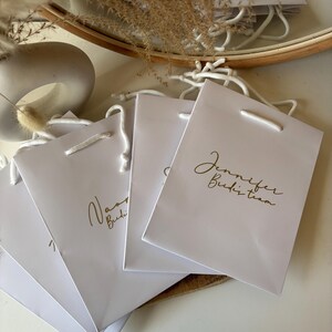 May include: White gift bags with gold lettering, featuring the text "Jennifer Budzi team" and "Naomi Budzi". Each bag has a white cord handle. The bags are arranged on a white surface.