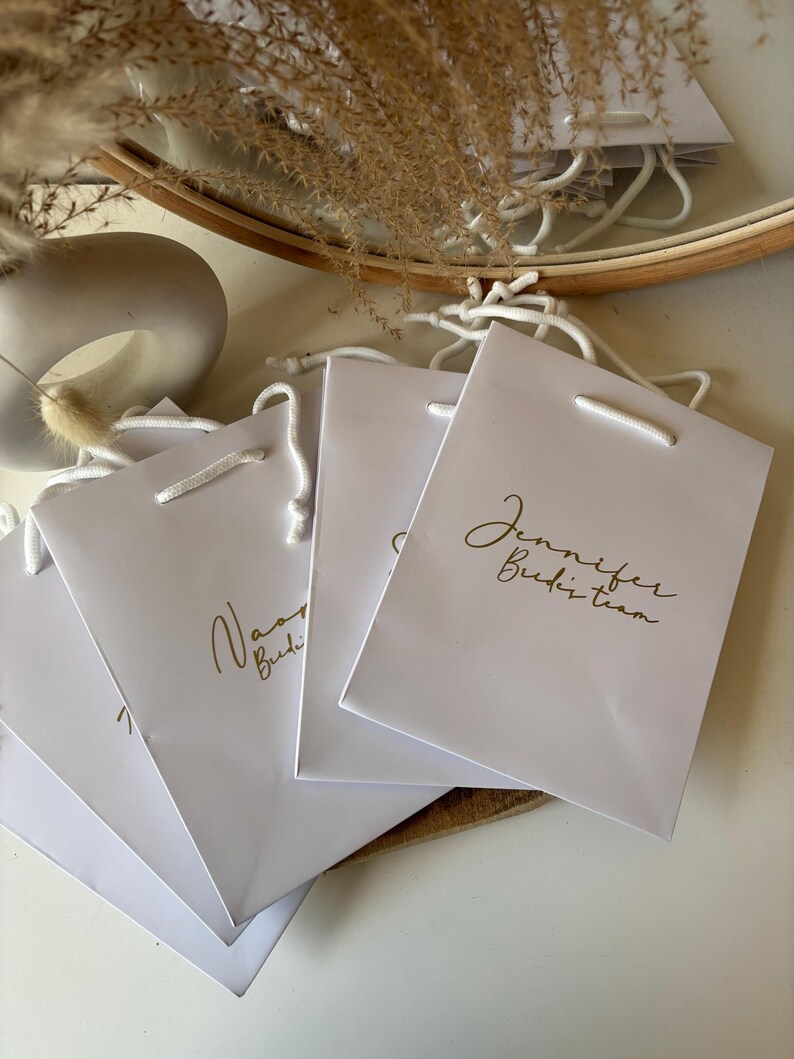May include: White gift bags with gold lettering, featuring the text "Jennifer Budzi team" and "Naomi Budzi". Each bag has a white cord handle. The bags are arranged on a white surface.