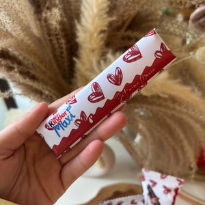 Peut inclure: Une barre de chocolat Kinder Maxi rouge et blanche avec un motif de cœurs et le texte "Je t'aime Marie".