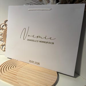May include: A white gift bag with a white cord handle. The bag has the name "No&eacute;mie" in a cursive font, with the text "Demoiselle d'Honneur en Or" below it. The date "30.05.2026" is printed at the bottom.