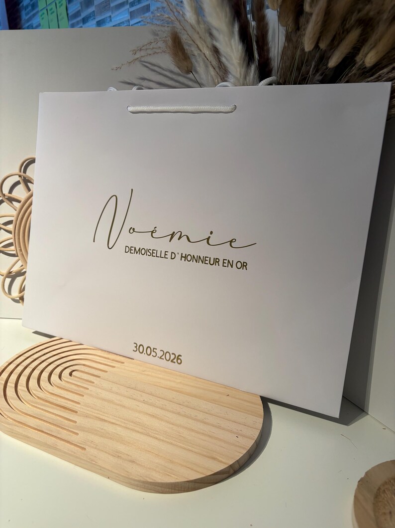 May include: A white gift bag with a white cord handle. The bag has the name "No&eacute;mie" in a cursive font, with the text "Demoiselle d'Honneur en Or" below it. The date "30.05.2026" is printed at the bottom.