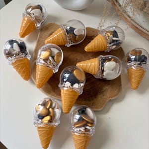 Puede incluir: Una colección de recipientes de plástico con forma de cono de helado llenos de caramelos variados. Los conos son de color naranja claro y las tapas transparentes contienen caramelos en tonos dorados, marrones y blancos. Los conos están colocados sobre una superficie de madera.