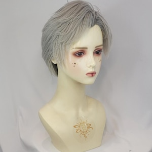 Puede incluir: Una cabeza de maniquí con cabello corto y degradado de color gris claro. El rostro presenta ojos pintados, cejas definidas y una boca pequeña. Un adhesivo dorado en forma de sol está centrado en el pecho y pequeñas estrellas doradas en las mejillas.