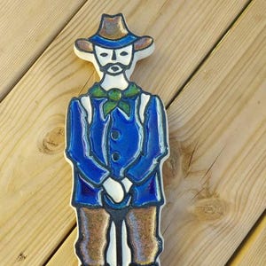 Puede incluir: Figura de cer&aacute;mica de un hombre con traje azul, pantal&oacute;n marr&oacute;n y sombrero, sosteniendo una pala. La figura tiene una cara blanca con bigote y barba. La pala tiene una hoja verde. Sobre una superficie de madera.