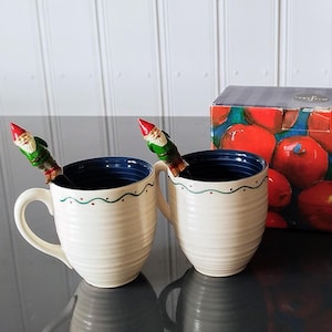 May include: Two white ceramic mugs with blue and green trim and a gnome-shaped spoon in each. The mugs are on a black surface and a box with red berries and the text "Mugg set" is in the background.