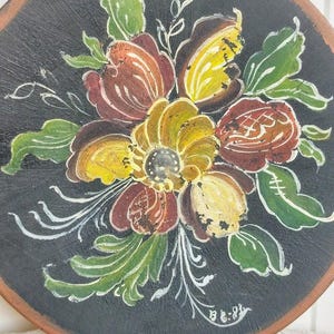 May include: Round, hand-painted wooden decorative piece featuring a floral design. The flowers are painted in shades of yellow, brown, and red, with green leaves and white accents, set against a black background. The edge is a natural wood color.