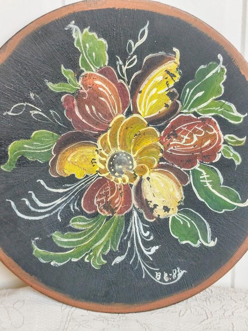 May include: Round, hand-painted wooden decorative piece featuring a floral design. The flowers are painted in shades of yellow, brown, and red, with green leaves and white accents, set against a black background. The edge is a natural wood color.